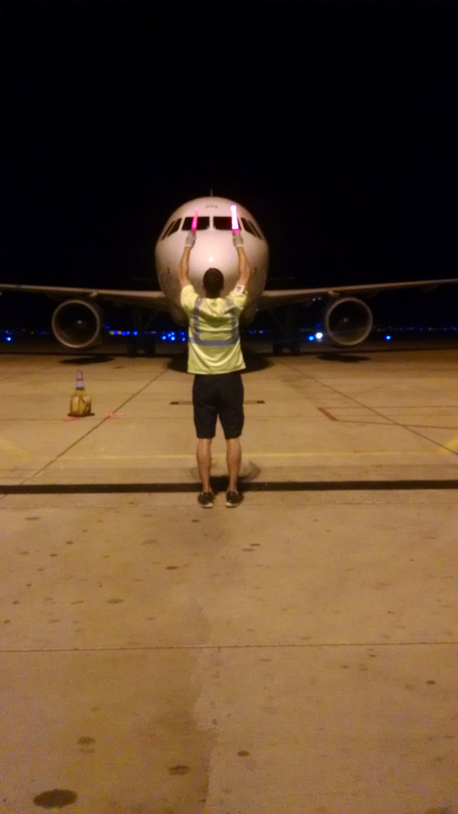Marshalling an arriving narrowbody airliner at night on the ramp, arms raised overhead with two lit wands, the aircraft nose stopped a few metres away
