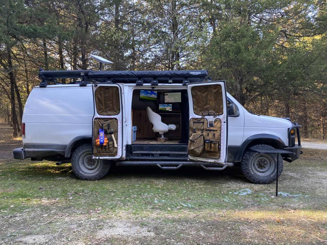 The van with both side doors open in a pine clearing, showing the interior desk, two monitors and a seat, with the awning extended overhead